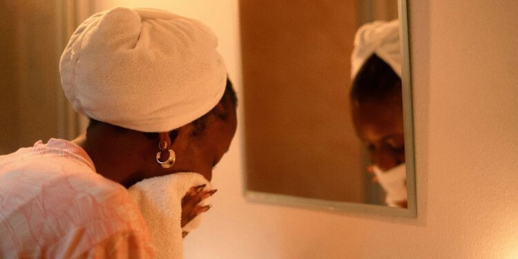 Woman with a towel wrapped around her hair drying her face in front of a mirror