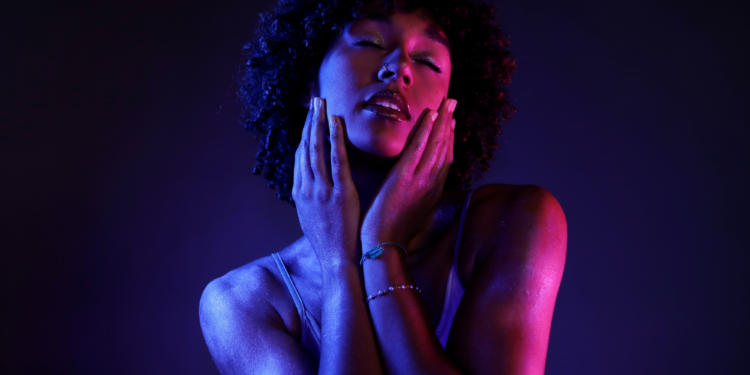 Close-up of a woman with curly hair, eyes closed, holding both hands to her cheeks under blue and pink lighting.