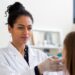 A clinician wearing a white coat and gloves examines a patient’s face during a consultation.