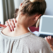 Woman seen from behind at a laptop, hand pressed to the back of her neck as she tilts her head forward.